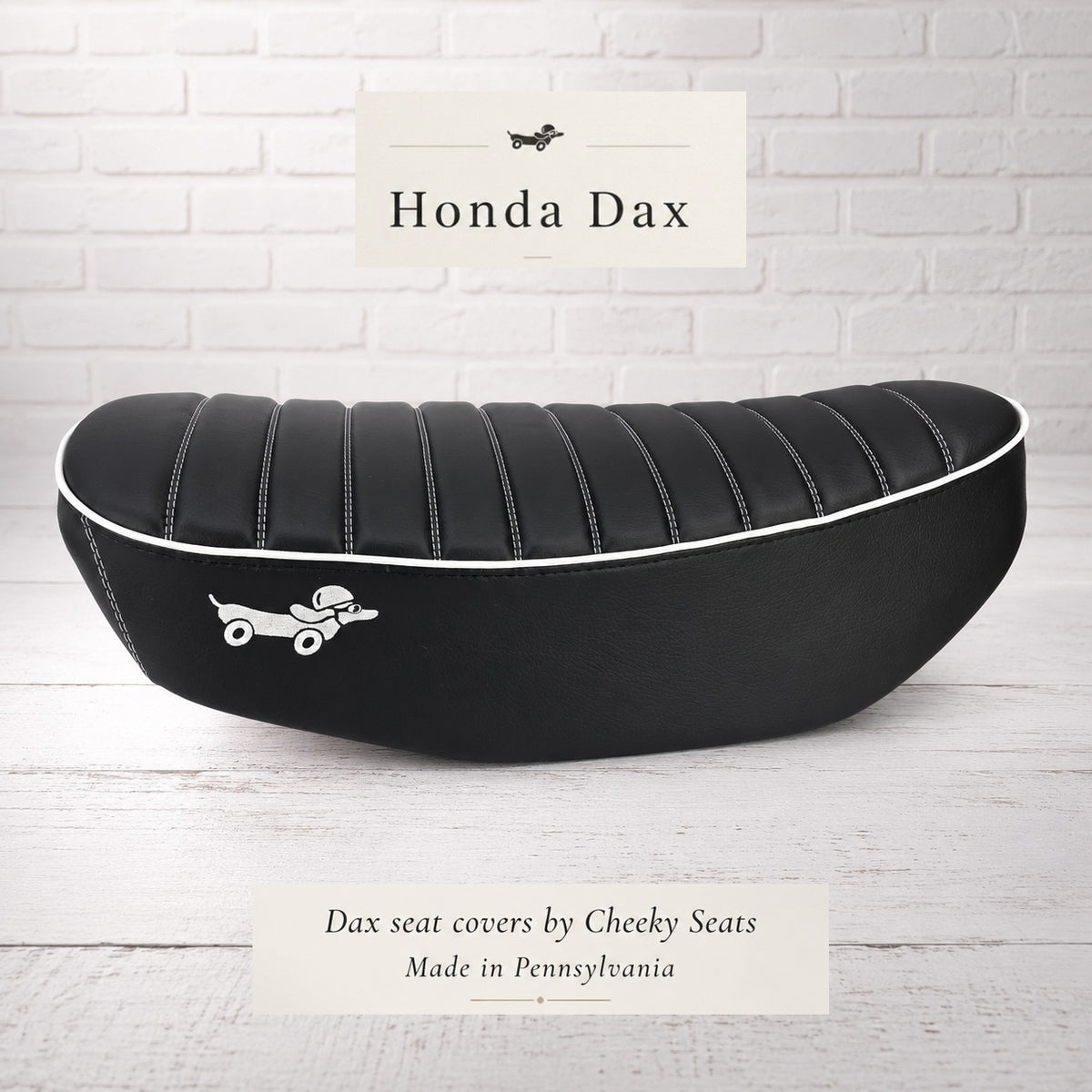 Black tuck-and-roll Honda DAX 125 seat cover with white piping and a playful Dax dog emblem, handmade in Pennsylvania by Cheeky Seats