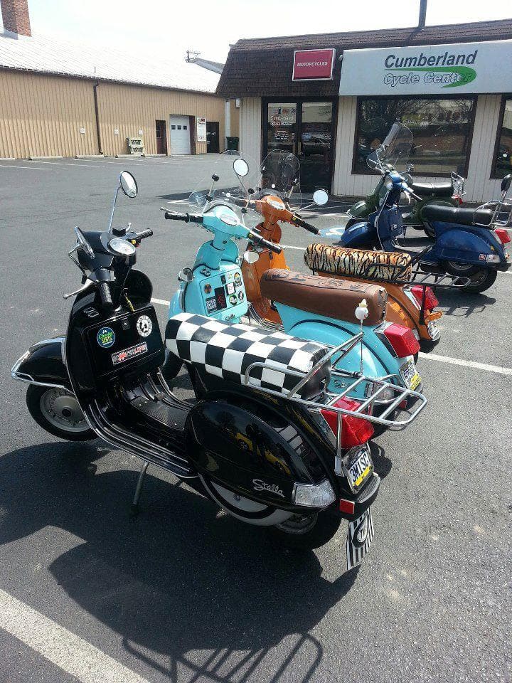 Black Genuine Stella with a handmade Cheeky Seats checker seat cover, parked among a colorful group of scooters