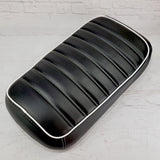 Black tuck-and-roll padded Maddog seat cover with horizontal pleats and white piping against a white brick backdrop, handmade by Cheeky Seats